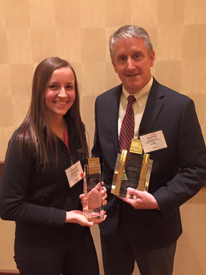 Man and woman standing while holding awards in front of a beige wall