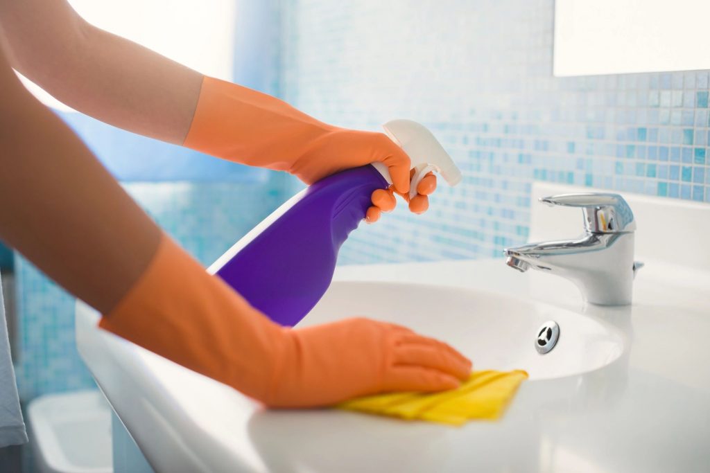 A close-up of a person with orange rubber gloves with spraying cleaner on a bathroom sink