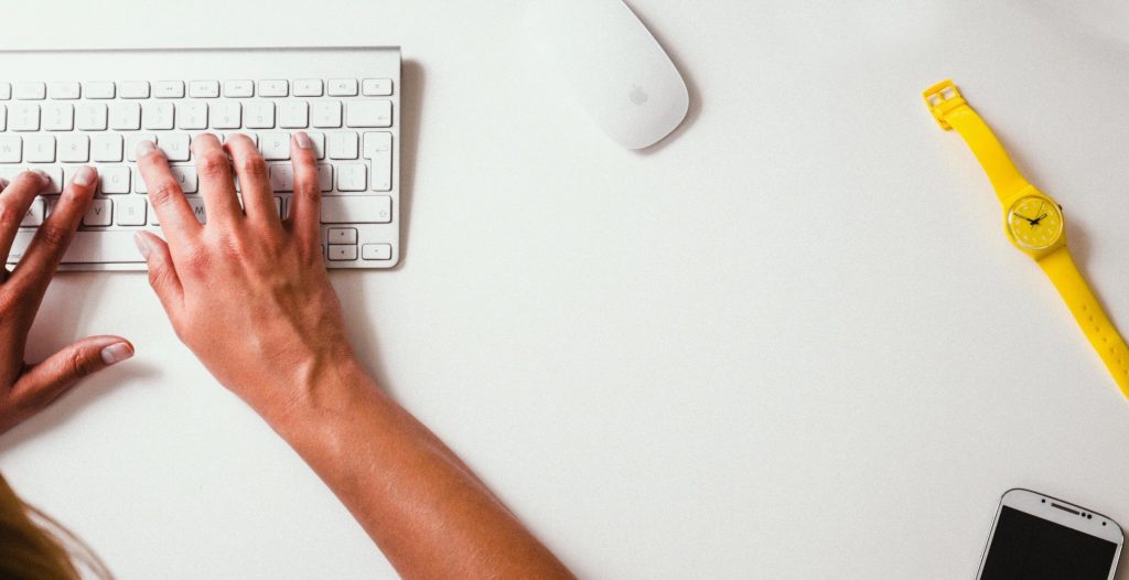 Overhead view of a pair of hands typing on a white keyboard on a white desk with a white mouse, yellow watch, and a mobile phone