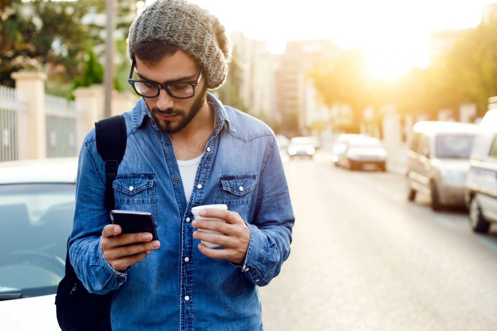 A man with a denim shirt and a knit hat looking at a cellphone and a small coffee cup