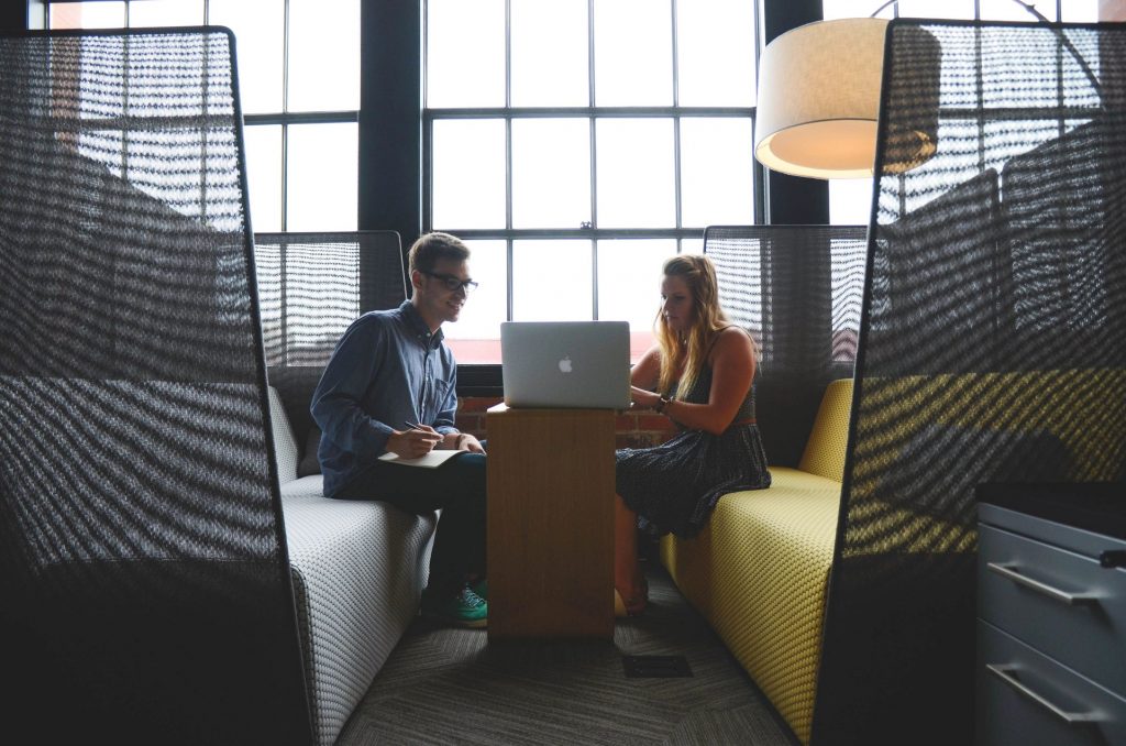 A young man and woman sitting on couches looking at laptop in front of a large window