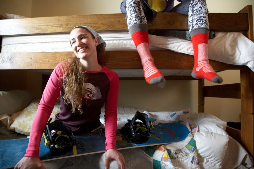 A girl with wearing a bandana sitting on a bottom bunk of a bunk bed with a snowboard across her lap