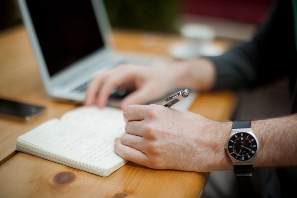 A close-up of a person writing in a notebook next to a laptop and a cellphone