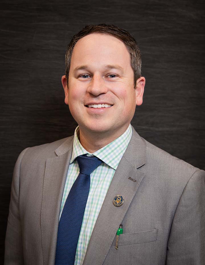 A man smiling for a professional picture with a grey jacket, green plaid shirt, and a blue tie