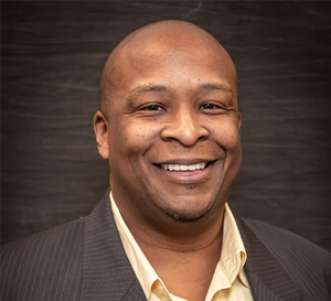 A man wearing a yellow shirt and brown jacket smiling for a professional photo