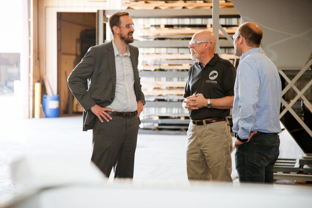 A man in a suit speaking with two men all wearing safety glasses