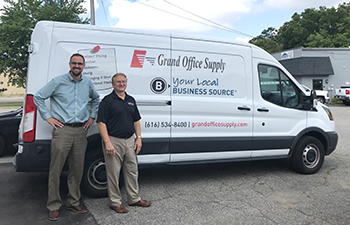 Two men posing for a shot outside of a Grand Office Supply delivery van