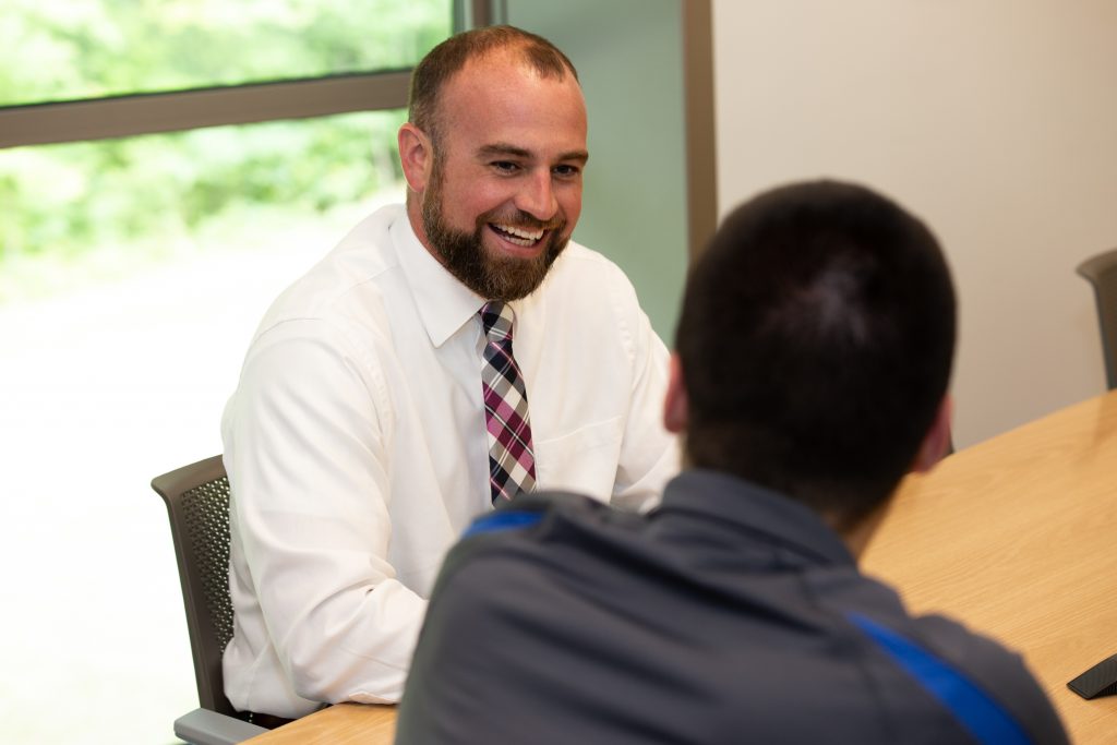 A man with a beard and a plaid tie smiling while speaking to a man at a conference table