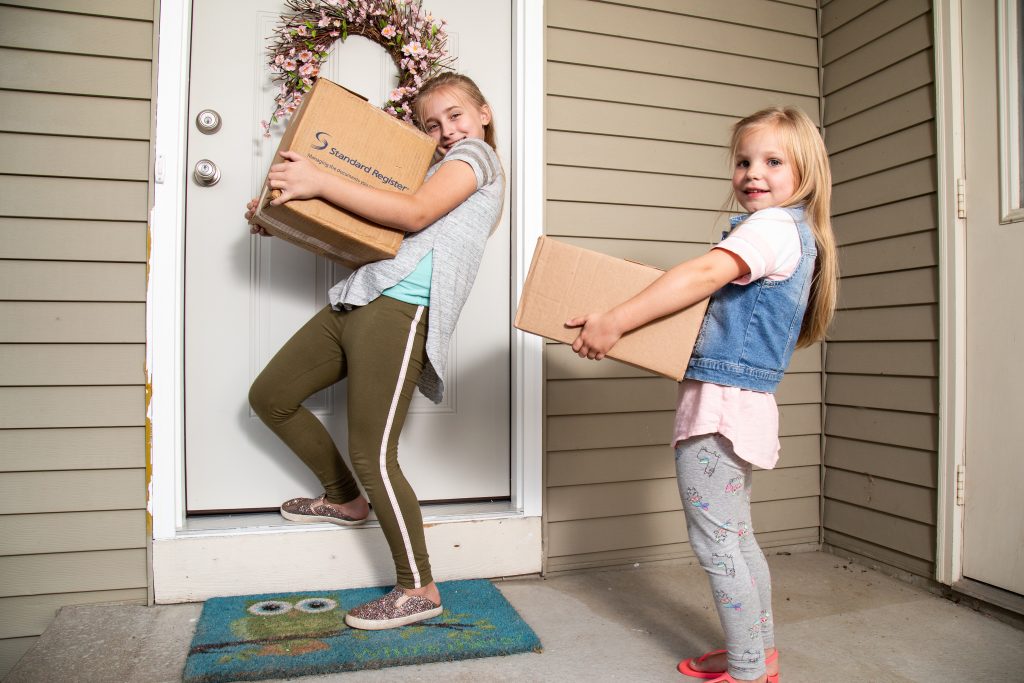 Two young girls holding cardboard boxes on a front porch about to enter the door