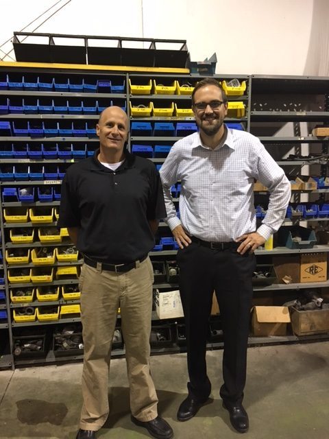 Two men smiling in front of shelves with blue and yellow containers in a workshop