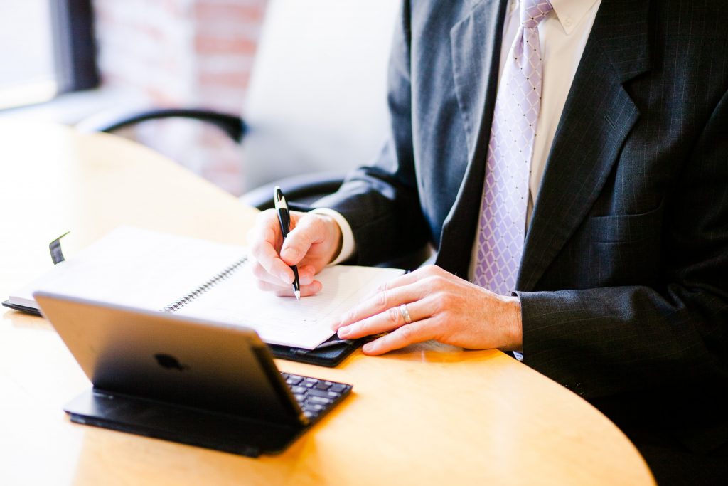 A man with a purple tie and a black suit writing in a planner in front of a mini iPad