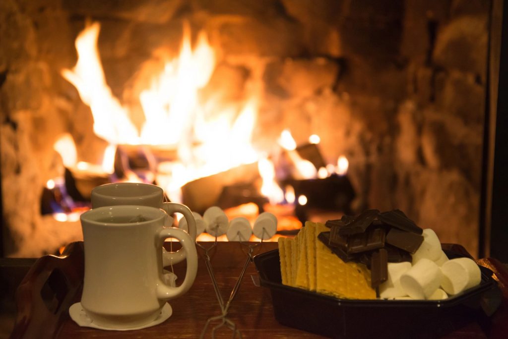 A two cups of tea, roasting sticks with marshmallows, and ingredients for s'mores in front of a fire