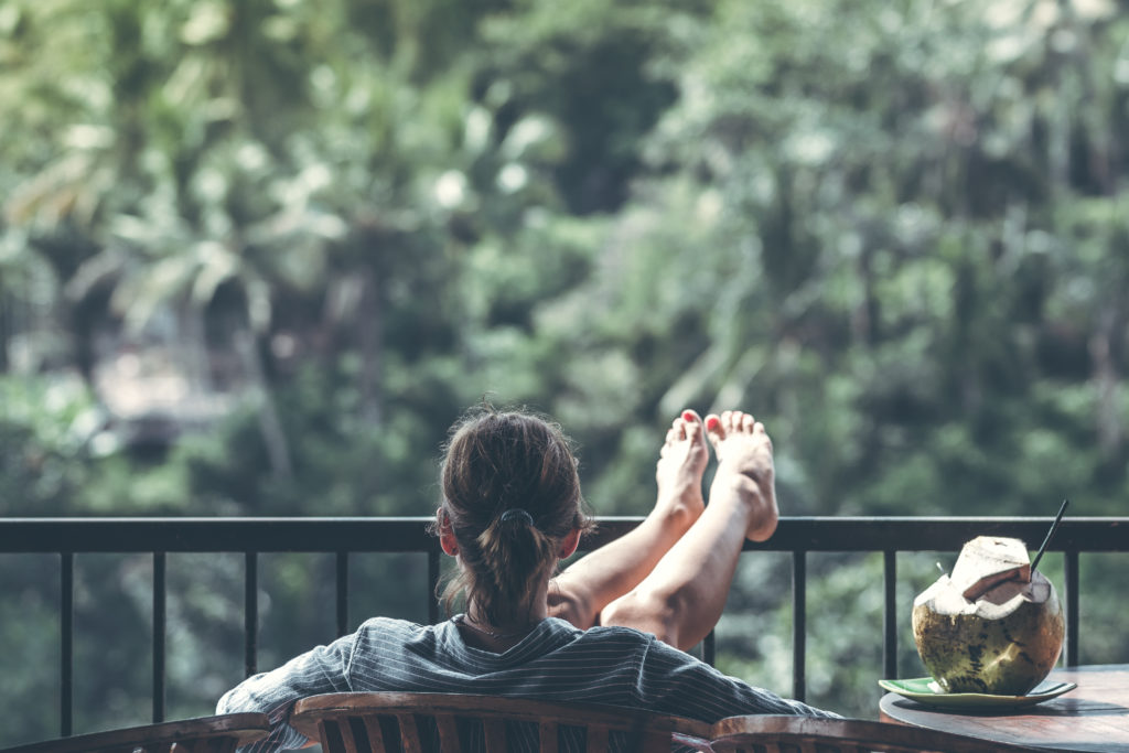 Woman sitting outside on brown wooden chair with feet up on railing beside a coconut